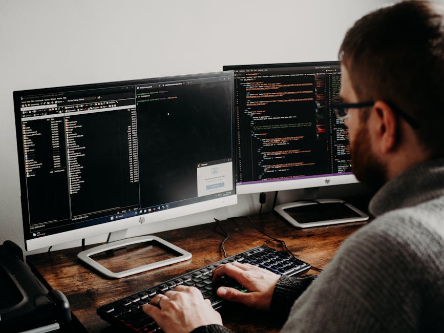 A man with glasses and a beard working at a wooden desk with two large computer monitors displaying lines of programming code on a black background. The left monitor shows a file directory and a text
